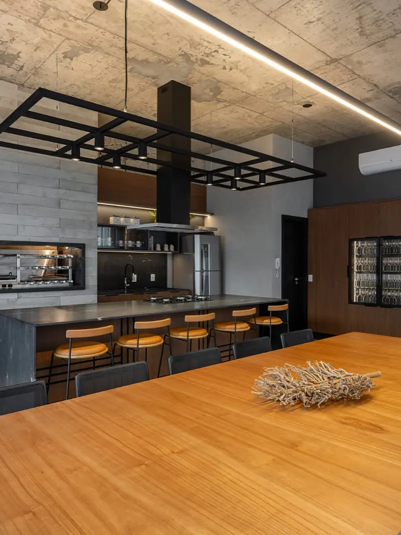 Espaço gourmet com concreto aparente, churrasqueira, ilha e banquetas.