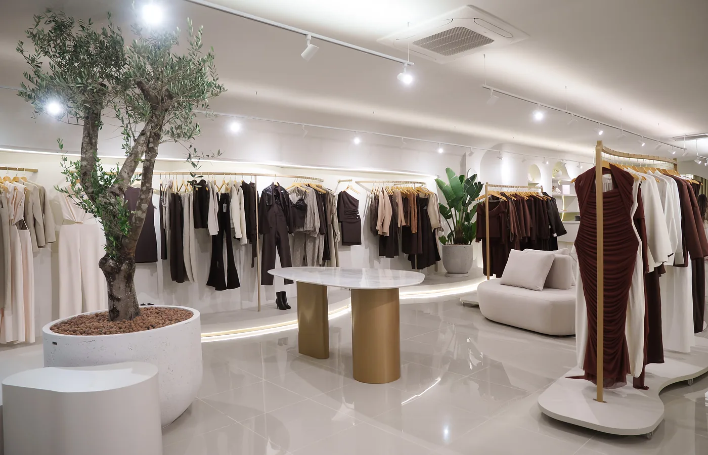 Loja de roupas com oliveira central, araras em latão e iluminação em trilho.