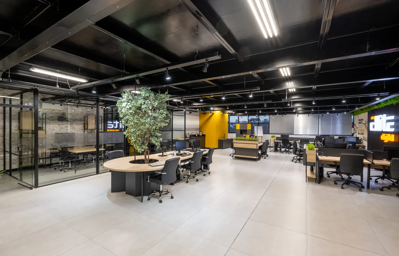 Escritório em planta aberta com estações de trabalho, salas de reunião envidraçadas e forro metálico preto.